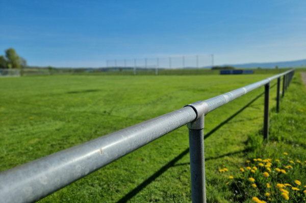 Rohrverbinder: Detailansicht eines verzinkten Metallgeländers mit einem deutlich sichtbaren Rohrverbinder vor einem weiten grünen Feld unter blauem Himmel.